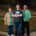 A teenage boy wearing a Shooting Sports National Championships shirt stands between his mother and father as they all smile. 