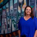 A smiling woman in a blue shirt stands in front of a wall mural.