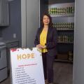 A professionally dressed woman standing in front of many rows of food cans and beside a sign that lists “HOPE.”