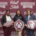 Three women standing in front of a maroon and white Mississippi State University banner.