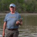 A man with a blue visor and T-shirt listing “Pearl Riverkeeper” and holding a clipboard in front of a waterway.