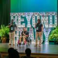 Three teens sing on stage, with the central performer seated in a wheelchair. Large, illuminated letters, arranged to form the words “Beyond Ready,” are in the background.