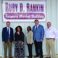 Five people, three men and two women, stand in front of a Farmers Market Building sign