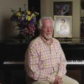 A male retiree in a plaid shirt and khaki slacks sits in front of a piano. On its soundboard is a picture of his late wife in her wedding gown and gloves.