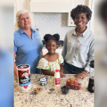 Two women and a child stand behind a kitchen counter filled with ingredients to make a smoothie