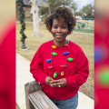 A woman stands on a sidewalk in a red sweatshirt and festive holiday-themed necklace.