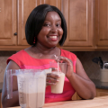 A woman holds a smoothie while standing in a kitchen.