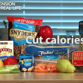 An assortment of snack foods on a butcher block table top, including a green pear, red grapes, baby carrots, pretzels, pecans, dates, almonds, and trail mix. 