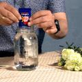 A man in a blue shirt adds a packet of flower food to a clear glass quart jar. White flowers lay beside the jar on a cloth-covered table.