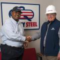 Two men shake hands in front of a Quality Steel sign.