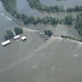 View from an airplane flying over extensive flood waters flowing over a levee and surrounding homes, farm buildings and crops.  