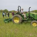 Man drives tractor pulling a large roller over a grass field while spectators watch.