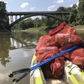 The front of a kayak has two red meshed bags containing litter and a blue trash-grabbing tool with the words “Pearl Riverkeeper” printed on it. The boat is on a small river and approaching a highway bridge.