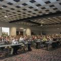 Hundreds of people in rows form an audience for a speaker in a large, open room.