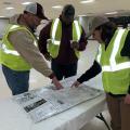 Two men and a woman, all wearing baseball caps and reflective yellow vests, look at a roadmap spread out on a table in a large, empty, well-lit room.