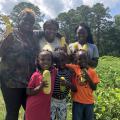 Two women and four children stand in a vegetable garden while holding yellow squash.