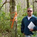 Man holding a clipboard looks at camera while standing in a wooded area beside a tree with orange ribbons tied around the trunk at shoulder height.