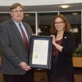 A man on the left and a woman on the right stand in an atrium and hold a framed document with a gold seal.