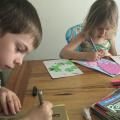 A boy and girl sit at a wooden table and use markers to draw on different colored sheets of paper.