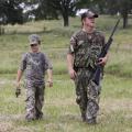 A man holding a shotgun and a boy dressed in camouflage walk in a grassy meadow.