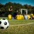 A soccer ball sits in the foreground with a soccer team and coach in the background.