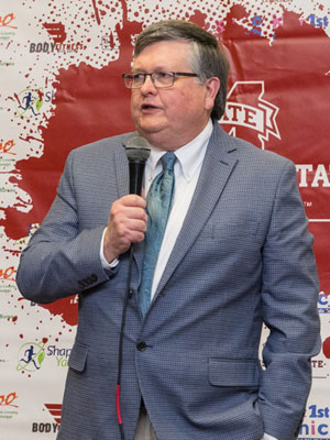 A white man standing, talking into a microphone.