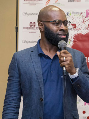 A Black man standing, talking into a microphone.